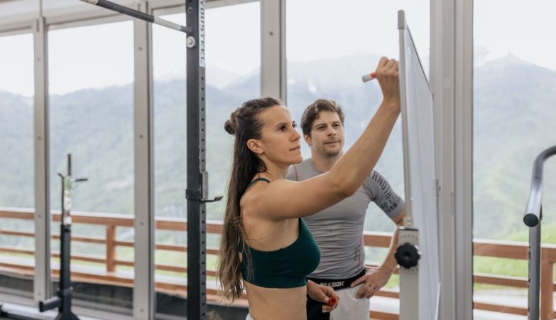 Female fitness coach writes on whiteboard as a male athlete watches in a gym.